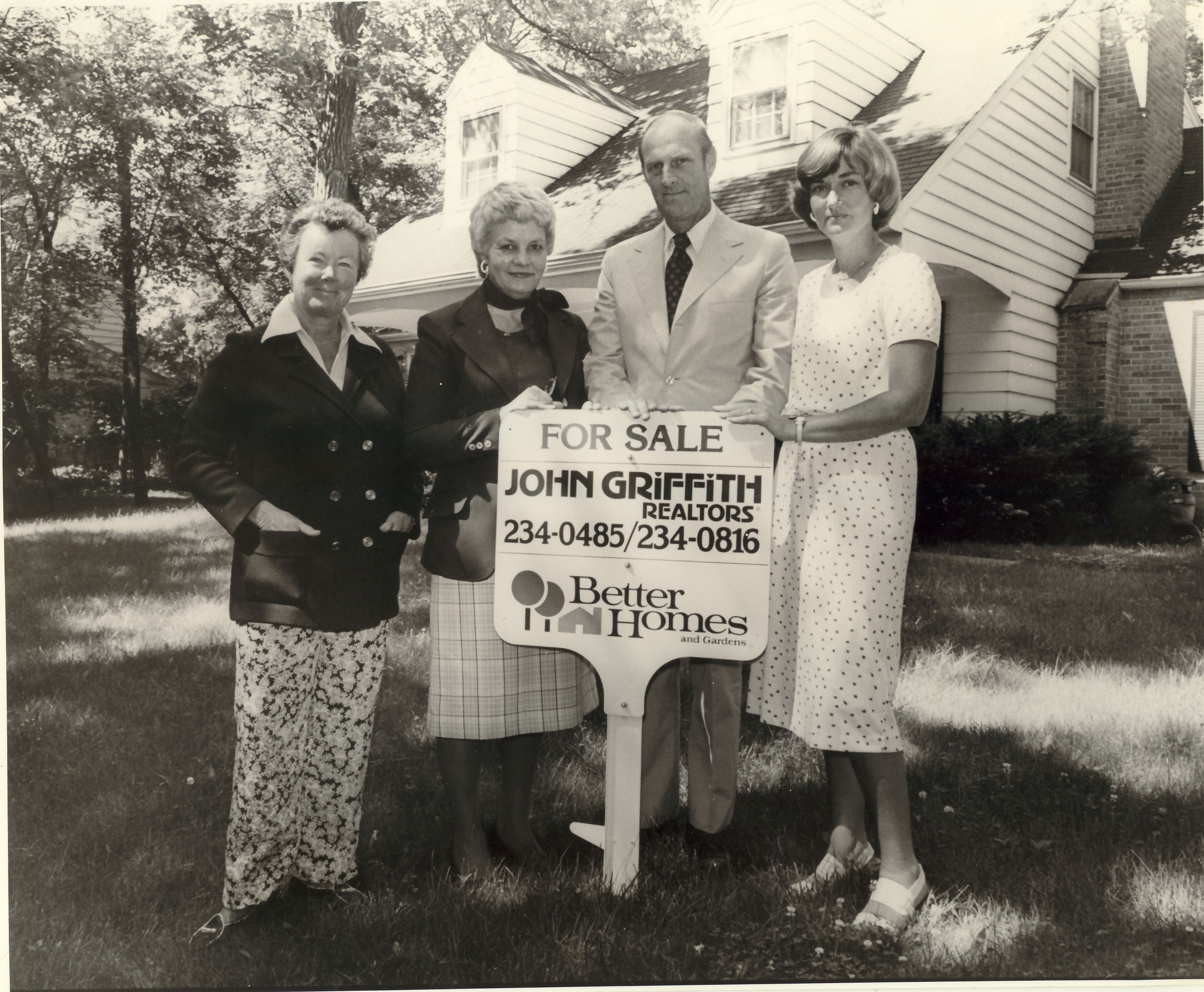John Griffith Realtors For Sale sign in front of unidentified home. Four realtors are standing beside the sign: Mary Griffis, Marjorie Carter, Gordon Lackie, and Susie Hoch. Home may be located at the northeast corner of South Green Bay Road and East Old Elm.