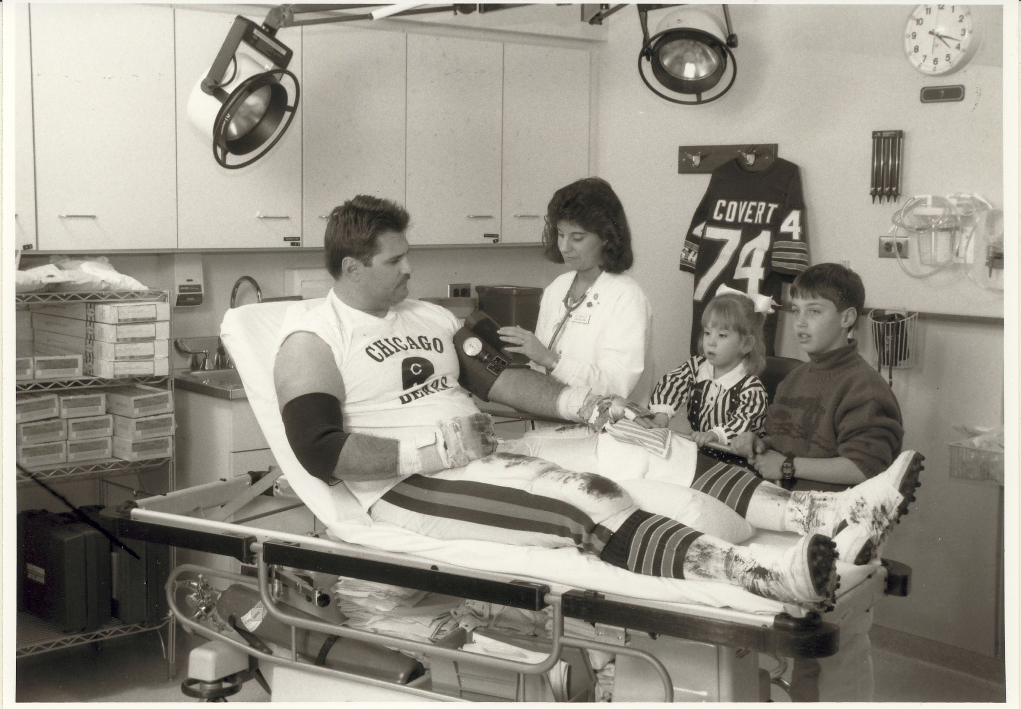 Chicago Bears offensive tackle Jim Covert is attended by Cathy Orr, R. N., at Lake Forest Hospital as his daughter and son look on, c. 1990. Covert is appearing in a special poster for nurse recruitment through the Illinois Hospital Association's “Nurses: Who Cares?” program.